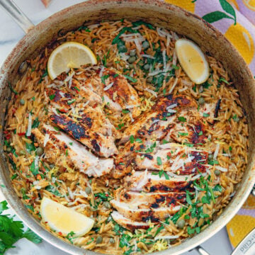 Closeup view of creamy lemon chicken orzo in a skillet with sliced chicken and lemon wedges over the top.