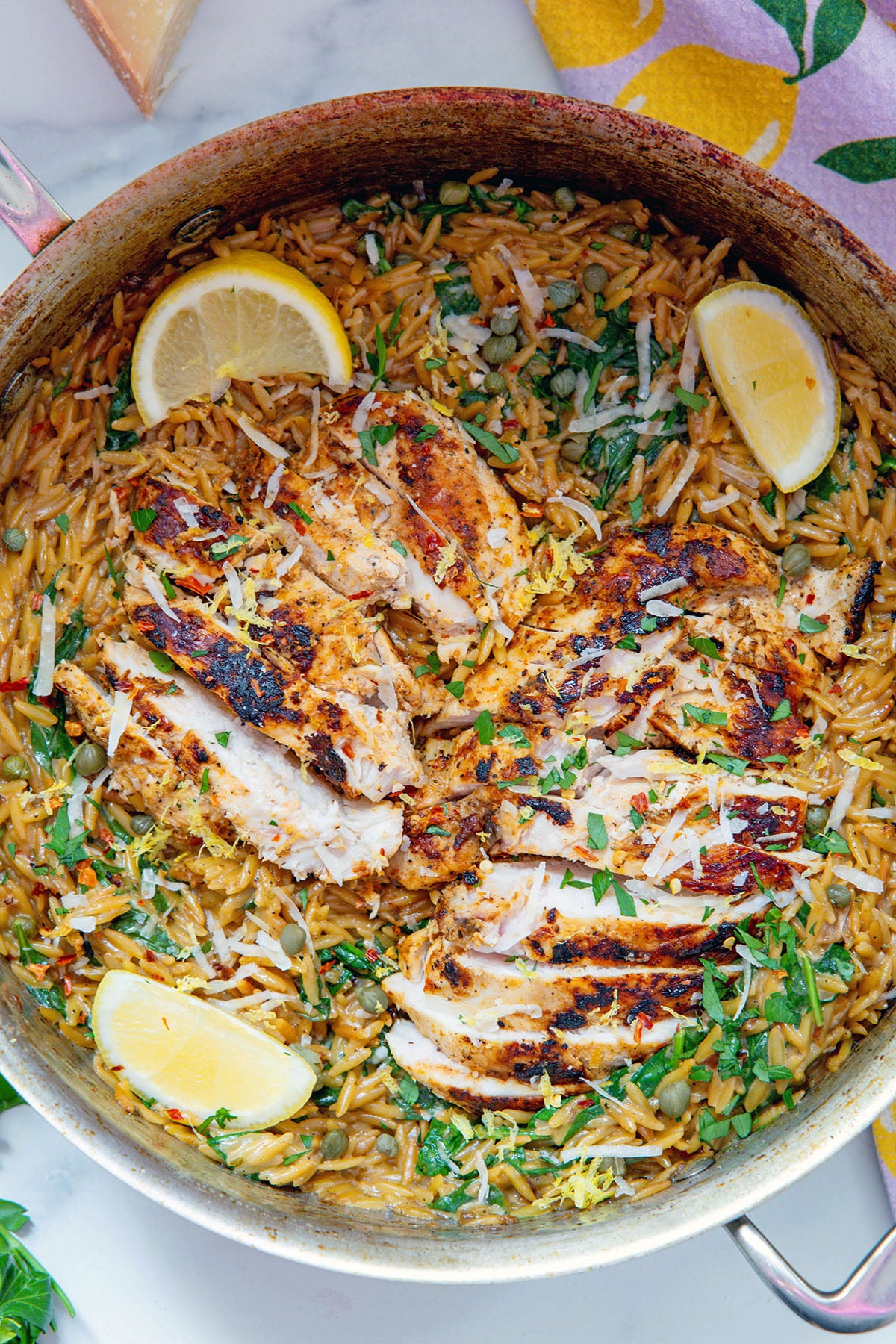 Overhead view of creamy lemon chicken orzo in a skillet with lemon wedges and chicken sliced along the top.