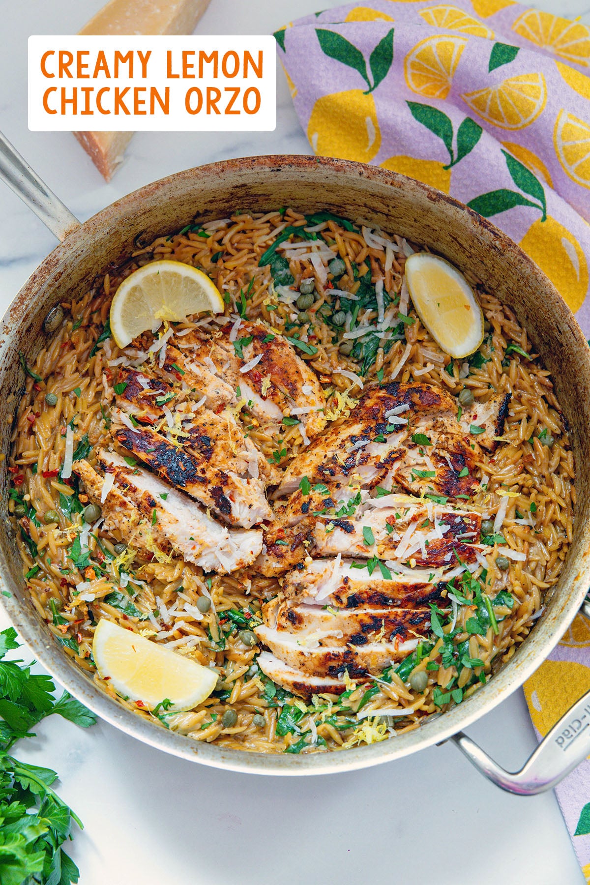 Overhead view of creamy lemon orzo in a stainless steel skillet with sliced chicken over the top and recipe title on photo.