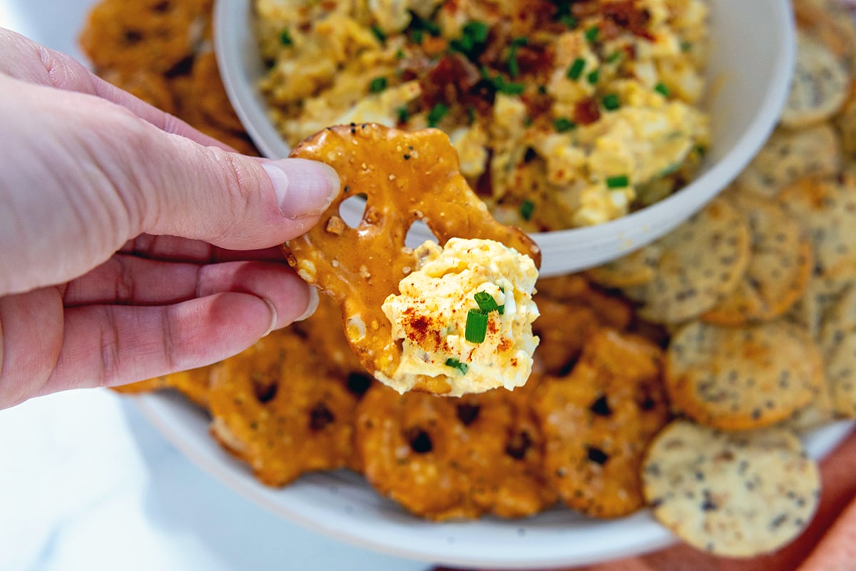 Deviled egg dip on a pretzel chip held over bowl.