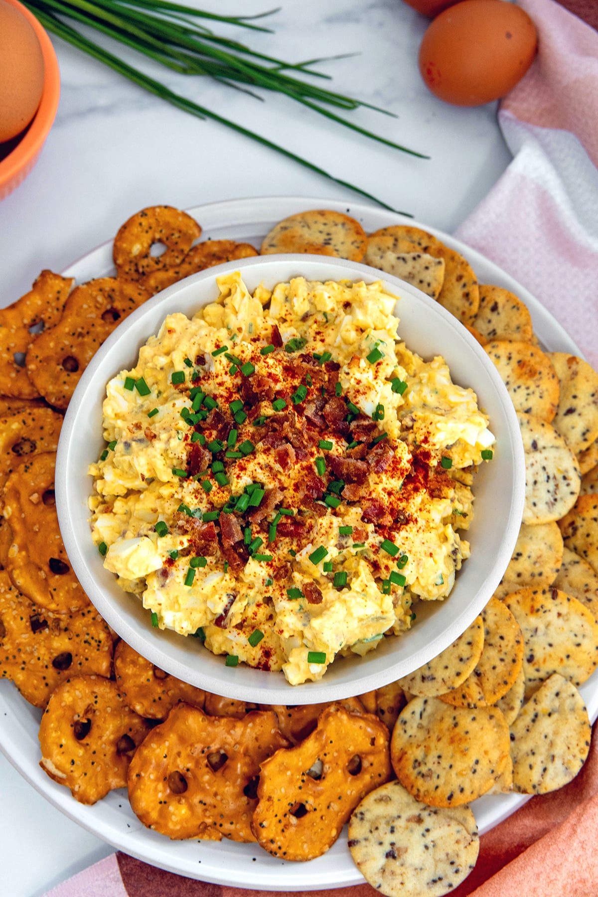 Overhead view of deviled egg dip in a bowl surrounded by pretzel chips and crackers with whole eggs and chives in the background.