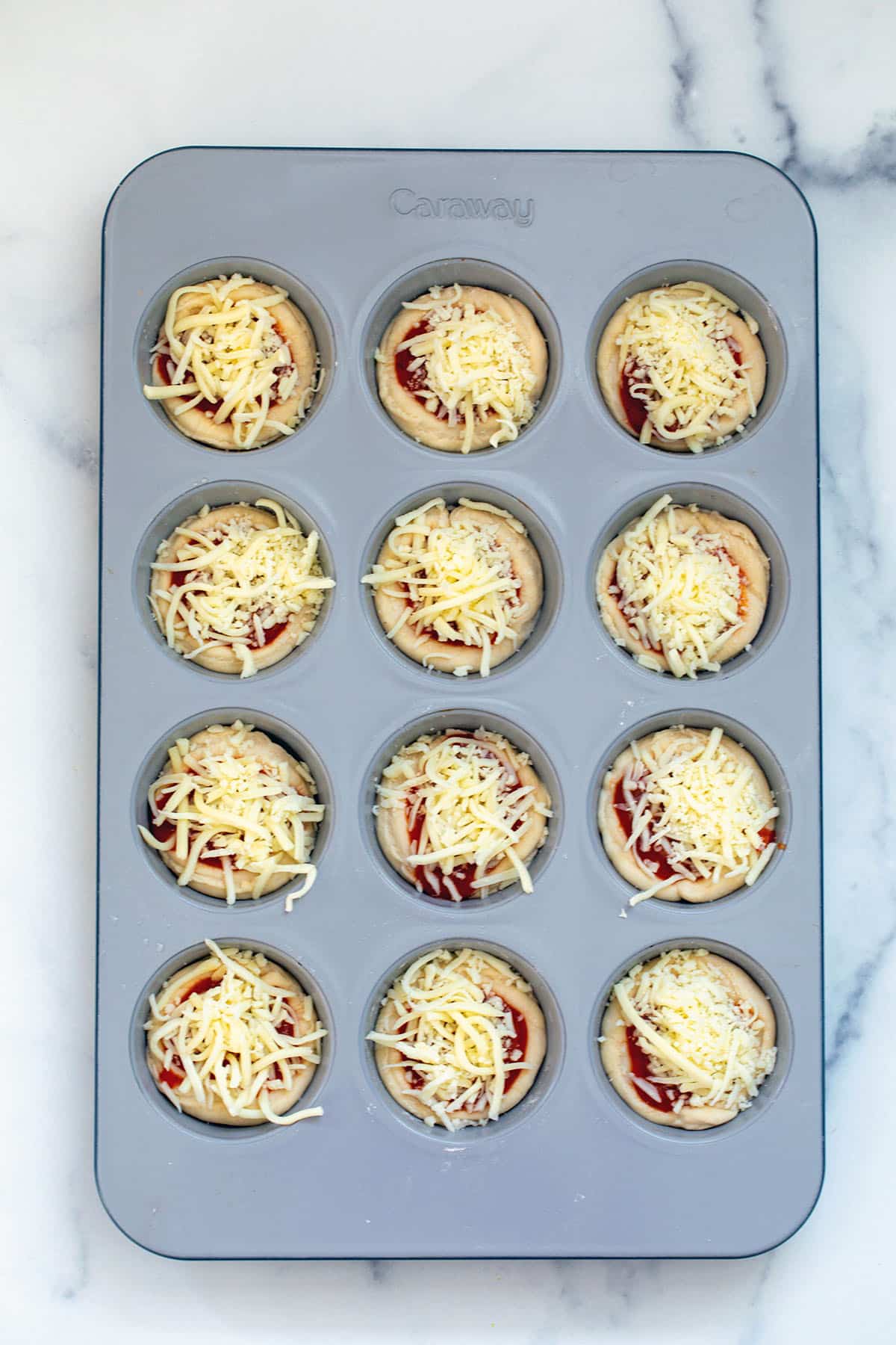 Cupcake tin with dough pressed into it and topped with sauce and shredded cheese.
