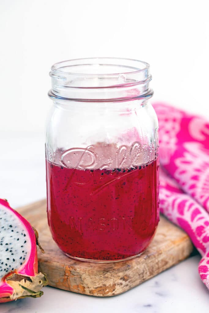 Head-on view of a mason jar of pink dragonfruit syrup with fresh dragonfruit half on the side.