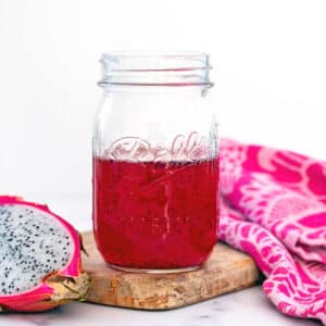 Mason jar of pink dragon fruit syrup with fresh dragon fruit half on side.