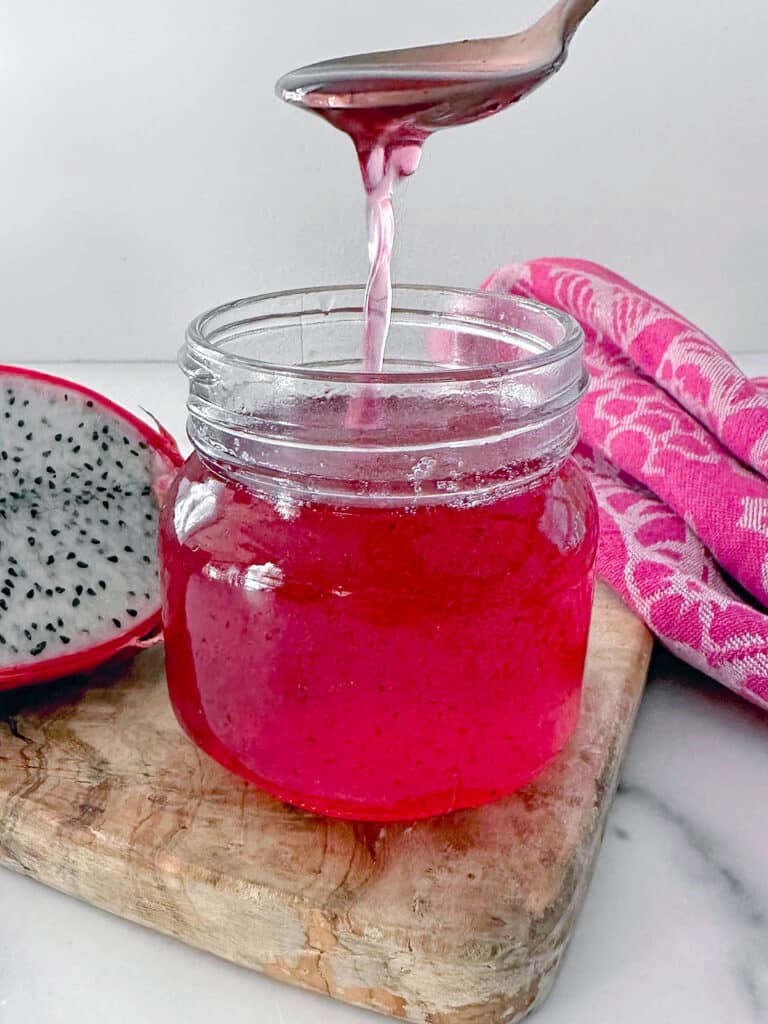 Pink dragon fruit syrup in a small jar with some being drizzled off a spoon with dragon fruit half in background.