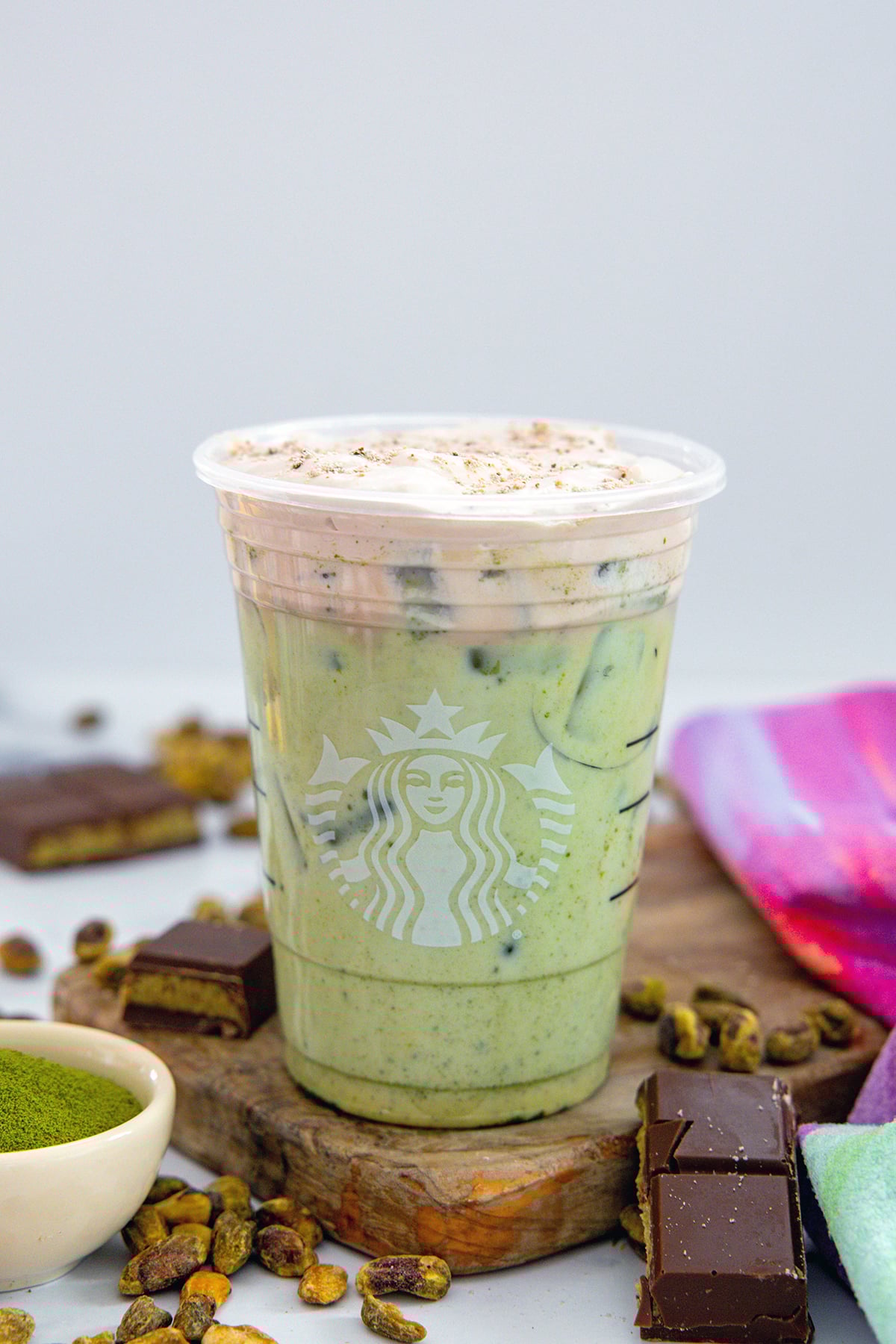 Head-on view of an Iced Dubai Chocolate Matcha with chocolate cold foam on top with small bowl of matcha, chocolate bars, and pistachios all around.