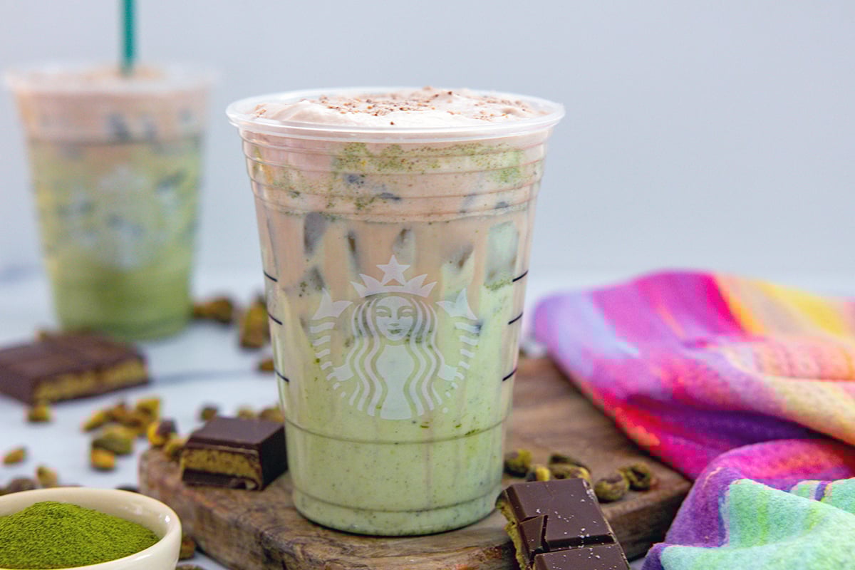 Landscape head-on view of two Dubai Chocolate Matchas with bowl of matcha powder and pistachios and chocolate bars all around.