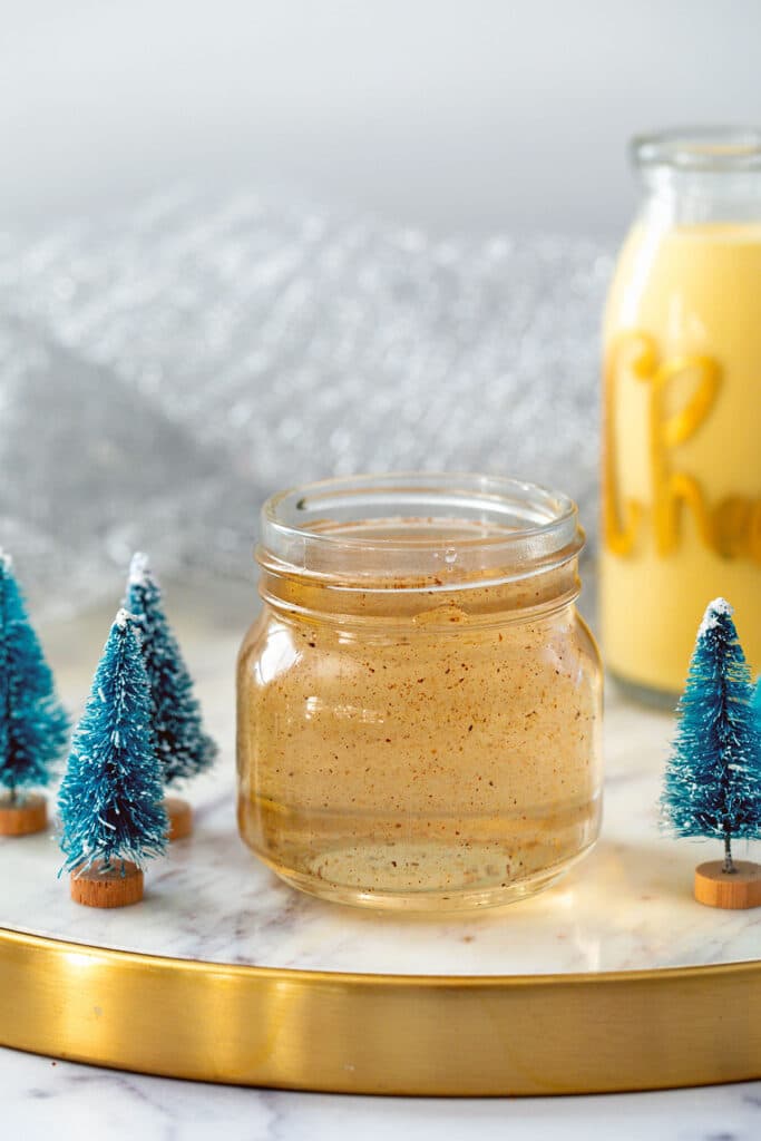 Small jar of eggnog syrup with bottle of eggnog and mini Christmas tree decorations all around.