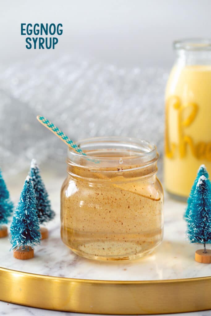 Small jar of eggnog syrup with glass of eggnog in background and small Christmas tree decorations.