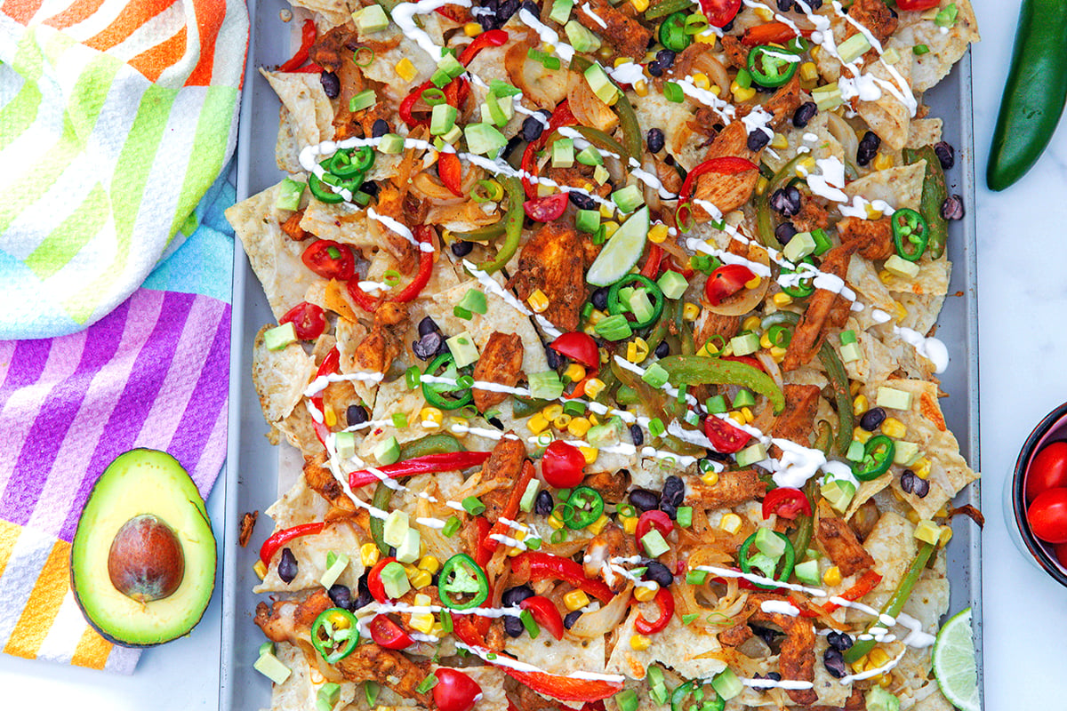 Landscape overhead view of chicken fajita nachos on a sheet pan with avocado half, jalapeño pepper, and tomatoes on the side.