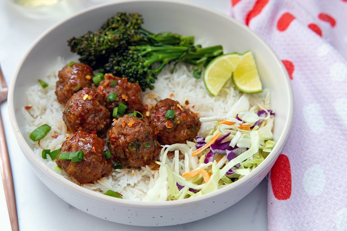 Landscape head-on view of a plate of spicy and sweet meatballs over coconut rice with slaw and broccolini.