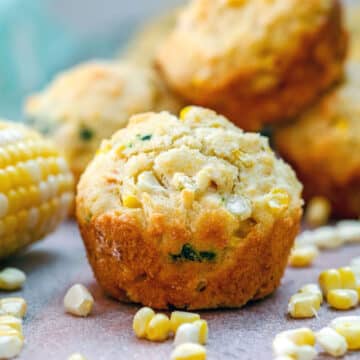 Closeup of a corn muffin with basil with corn kernels and corn on the cob all around.
