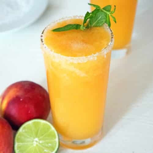 Overhead closeup view of a frozen peach margarita with lime wedge and peaches in background.