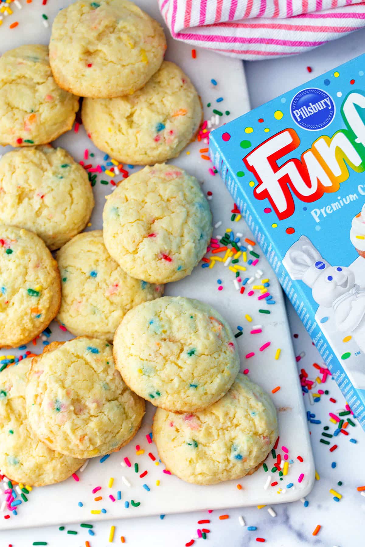 Funfetti cake mix cookies on a platter with a box. of cake mix next to them with rainbow sprinkles all around.