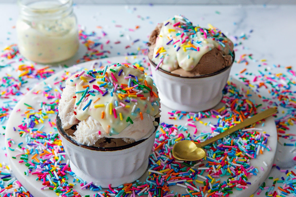 Landscape head-on view of two bowls of ice cream topped with funfetti magic shell with sprinkles all around.