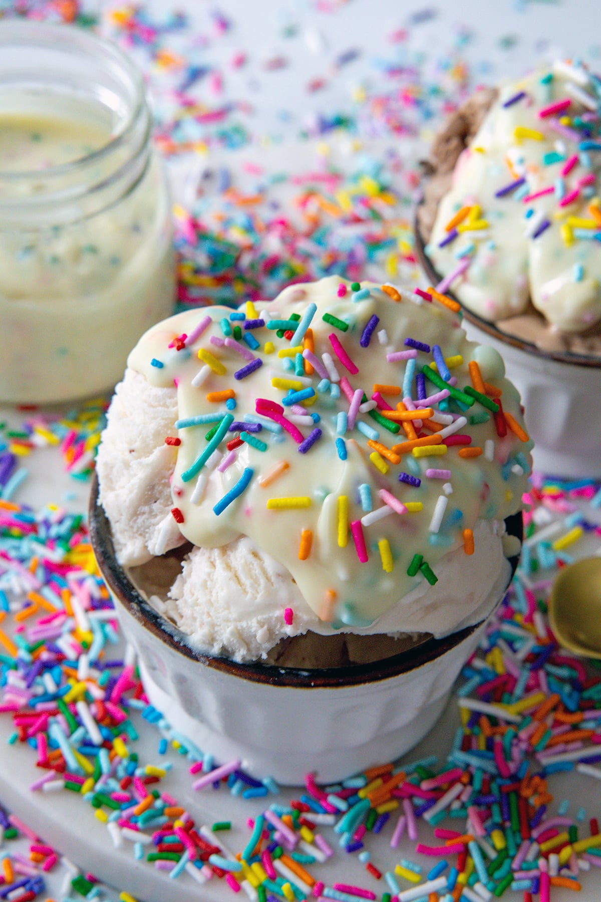 Scoop of ice cream topped with funfetti magic shell with sprinkles with second bowl and jar of magic shell in background.