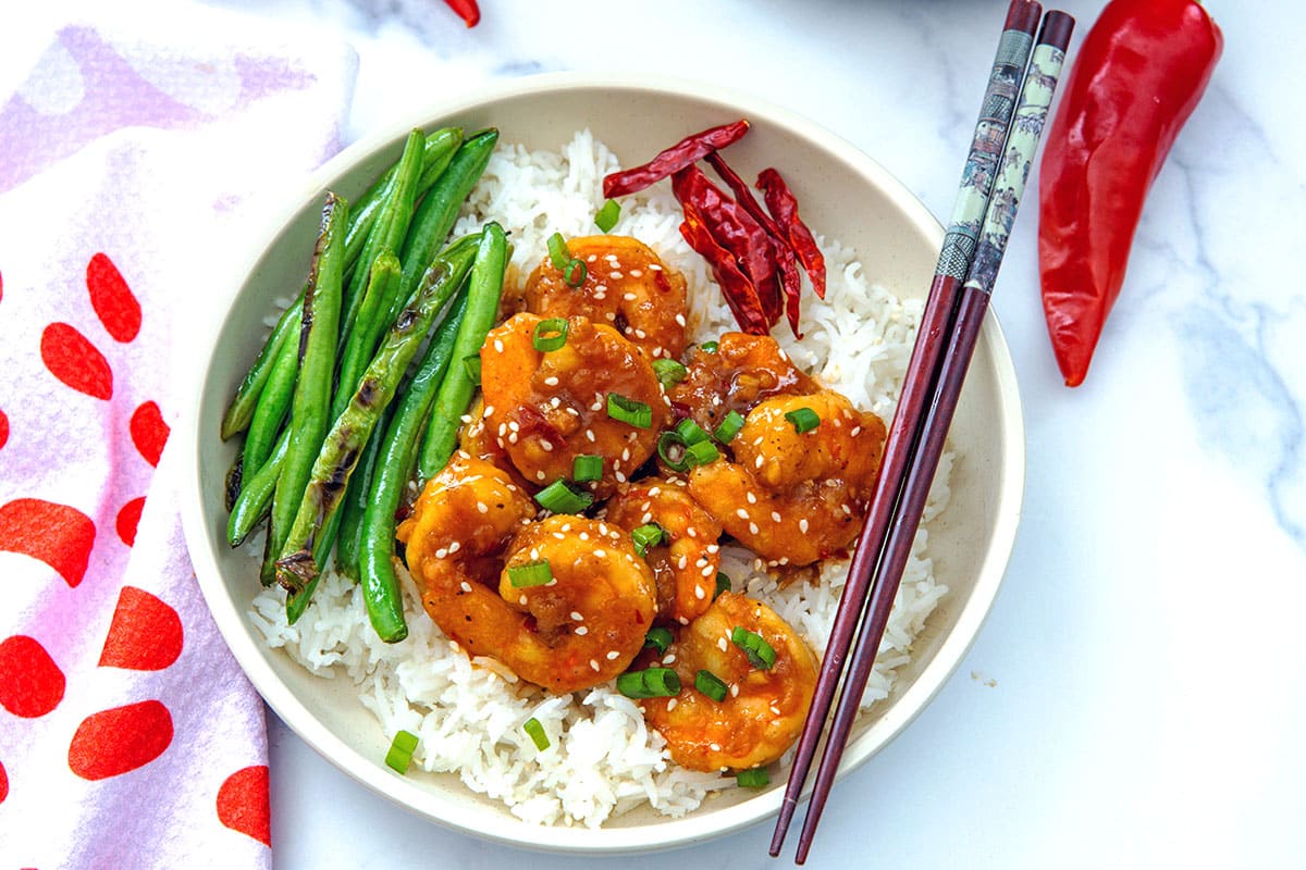 Landscape overhead view of a bowl of General Tso's shrimp over white rice with green beans with chopsticks and red chili peppers.