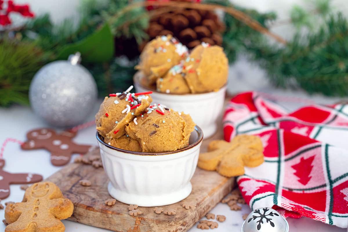 Landscape head-on view of two bowls of gingerbread cookie dough with sprinkles all around and gingerbread men, ornaments, and garland in background.