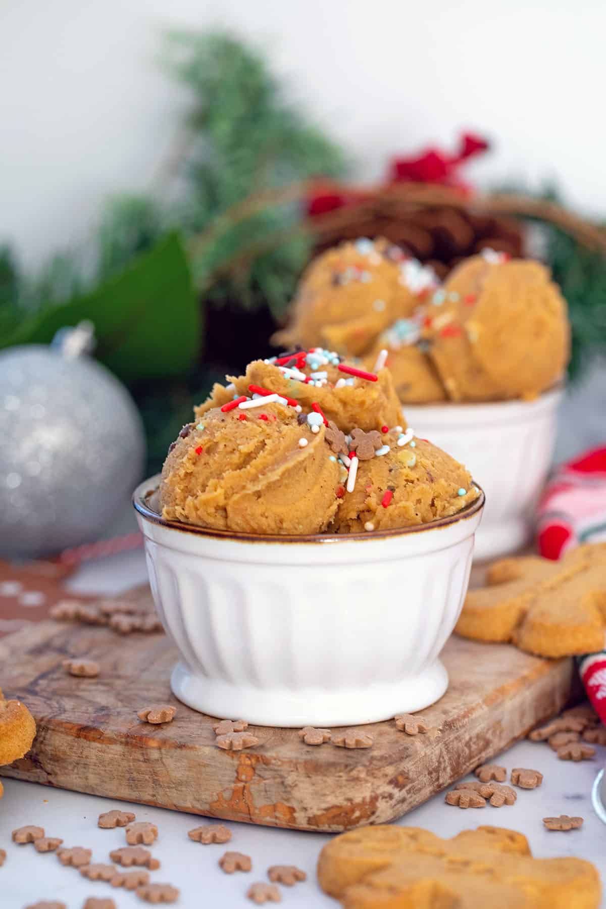 Head-on view of gingerbread cookie dough in small bowls with sprinkles and holiday garland in background.