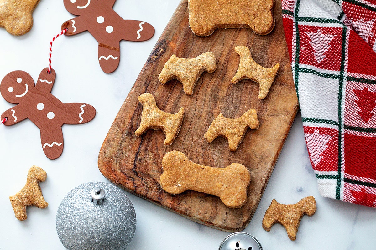 Landscape overhead view of dog and bone-shaped gingerbread cookies for dogs on a wooden board with silver ornaments and gingerbread men decorations in background.