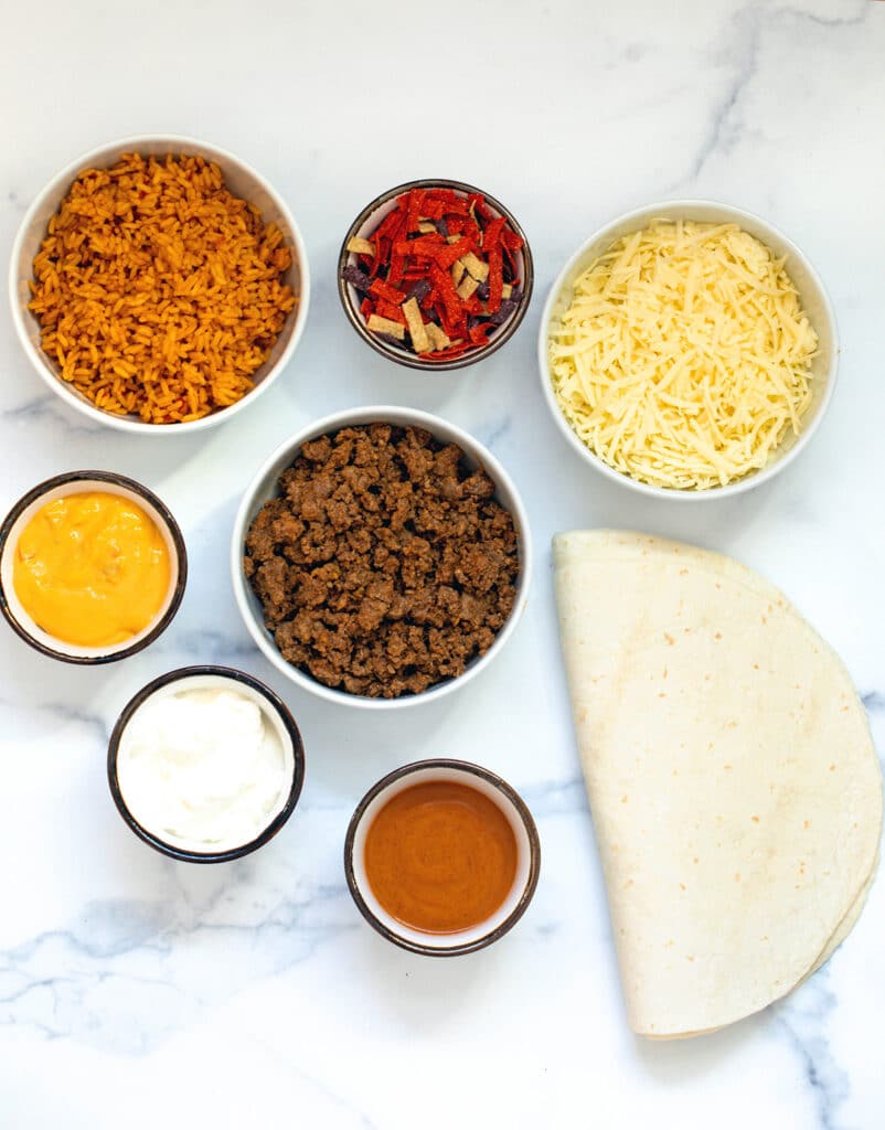Ingredients for grilled cheese burritos, including seasoned beef, seasoned rice, shredded cheese, sour cream, chipotle sauce, queso, fiesta strips, and tortillas.