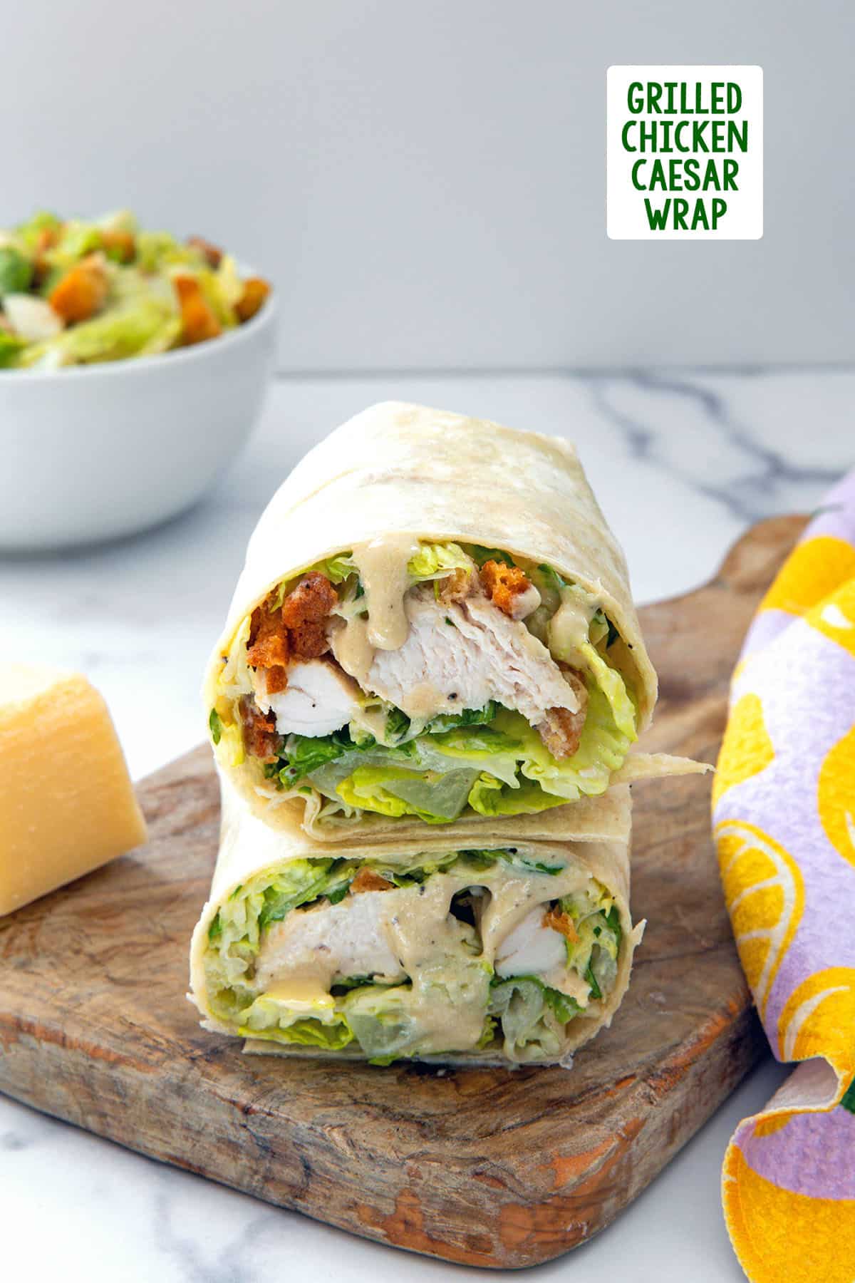 Two chicken caesar wraps stacked on each other with bowl of salad in background, block of parmesan on side, and recipe title at top.