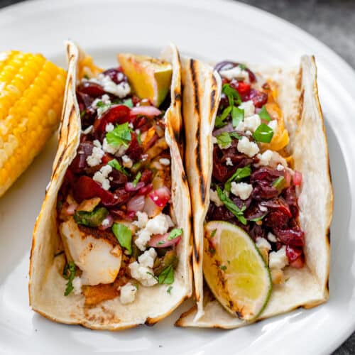 Closeup view of two flour tortilla tacos with cod and cherry salsa and a lime wedge.