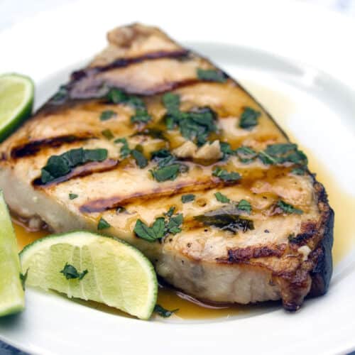 Closeup view of mojito swordfish with grill marks on it, topped with chopped mint with sliced limes on the side.