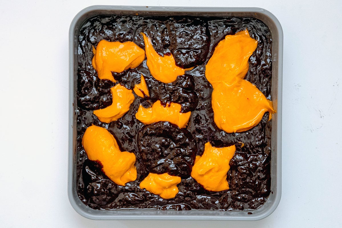 Brownie batter in pan with orange cream cheese mixture dolloped on top.