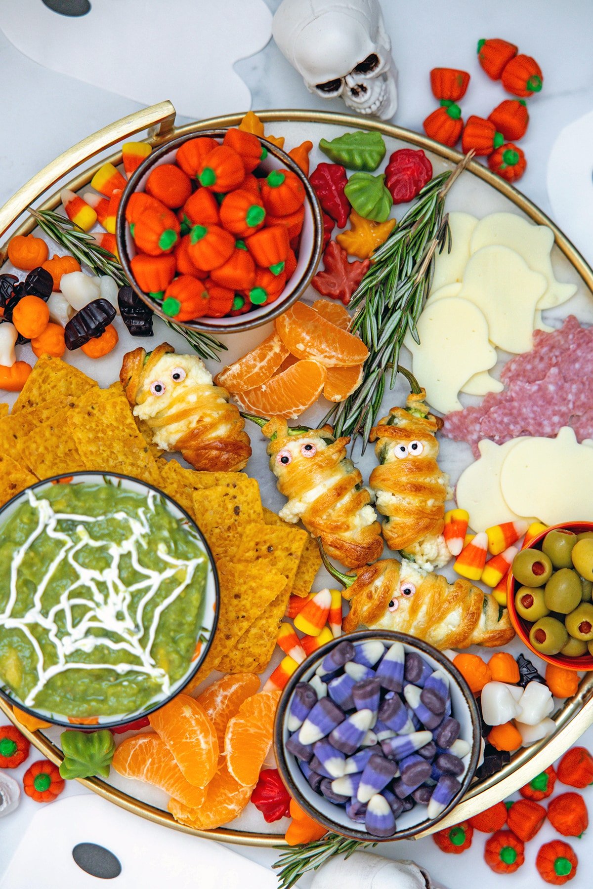 Overhead view of Halloween charcuterie board with candy corn and other festive candies, tortilla chips, jalapeño popper mummies, spiderweb guacamole, and more.