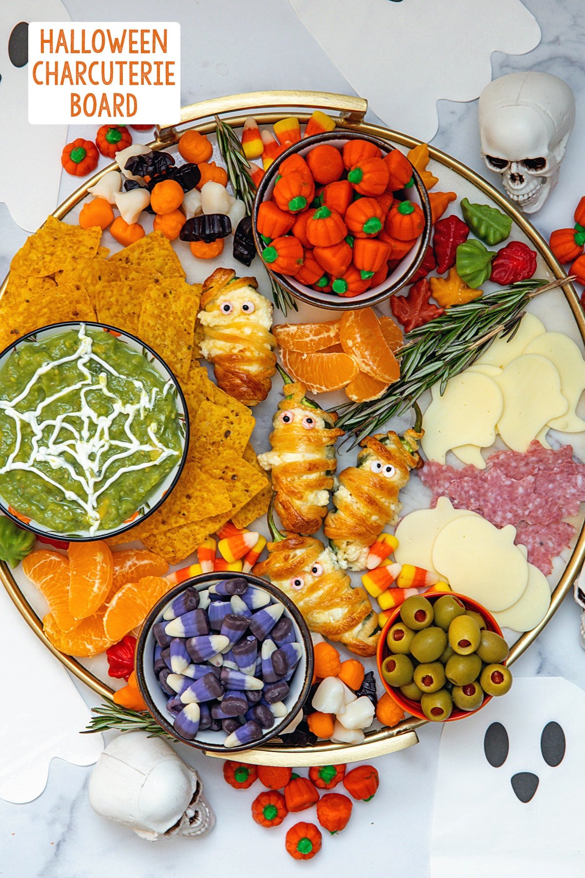 Overhead view of a Halloween charcuterie board with jalapeño popper mummies, spiderweb guacamole, candy corn, ghost and pumpkin cheese and salami, tortilla chips, olives, and candies with "Halloween Charcuterie Board" text at top.