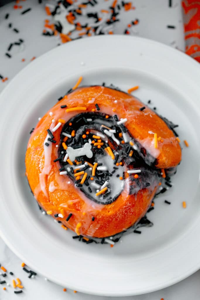 Overhead view of an orange Halloween cinnamon roll on a plate with sprinkles all around.