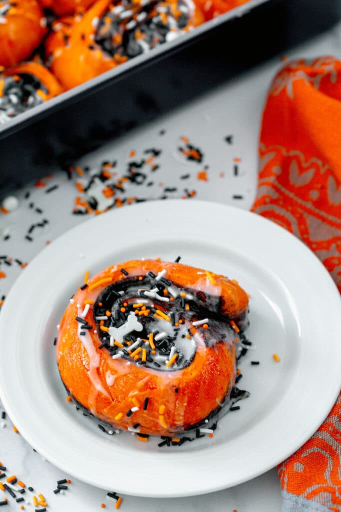 Orange Halloween cinnamon bun with chocolate filling, icing, and sprinkles on white plate with pan of cinnamon buns in background.