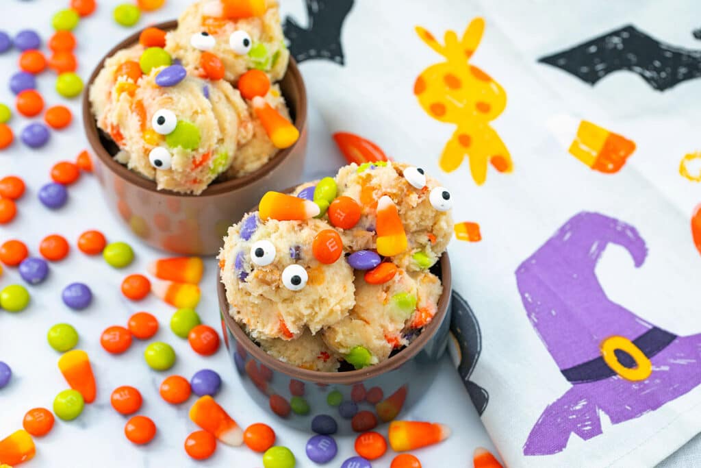 Landscape overhead view of two bowls of Halloween cookie dough with festive. M&Ms, candy corn, and Google eyes.