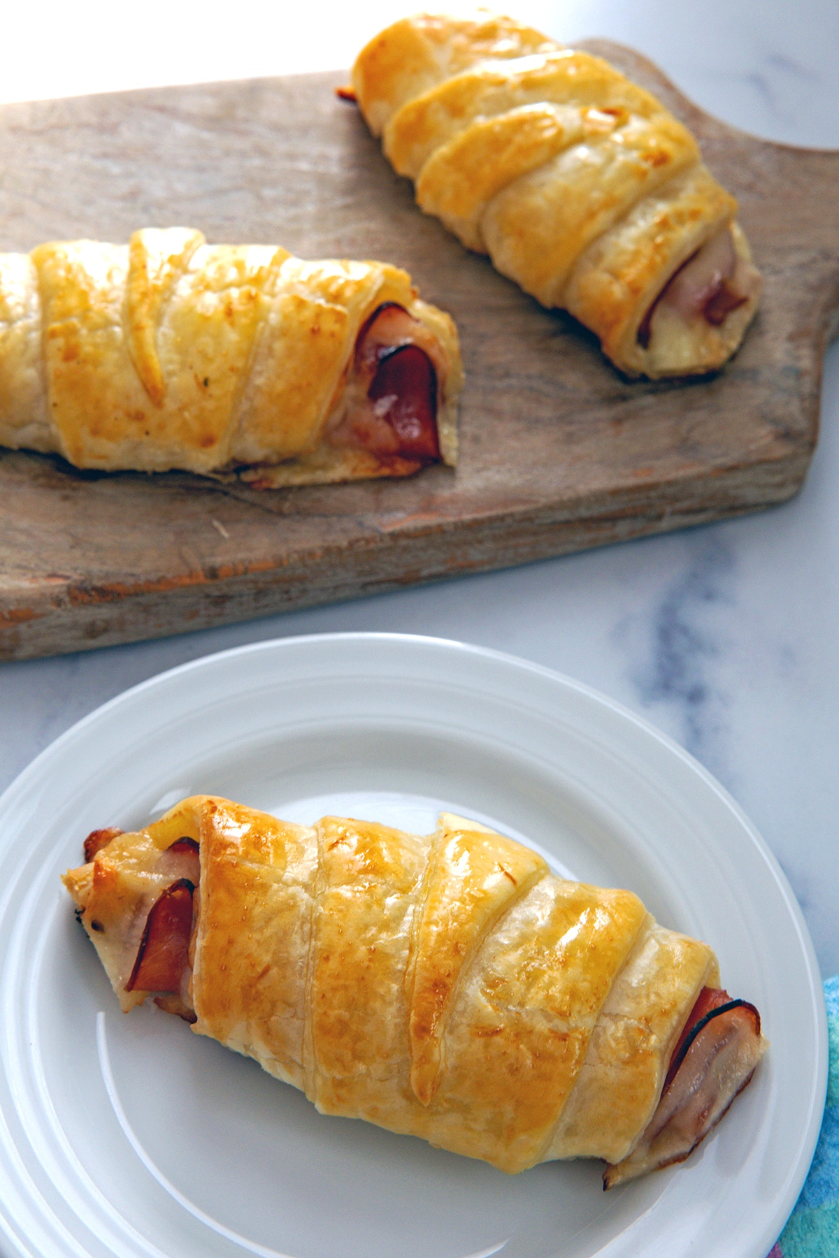 Ham and cheese croissant on a white plate with two more on board in background.