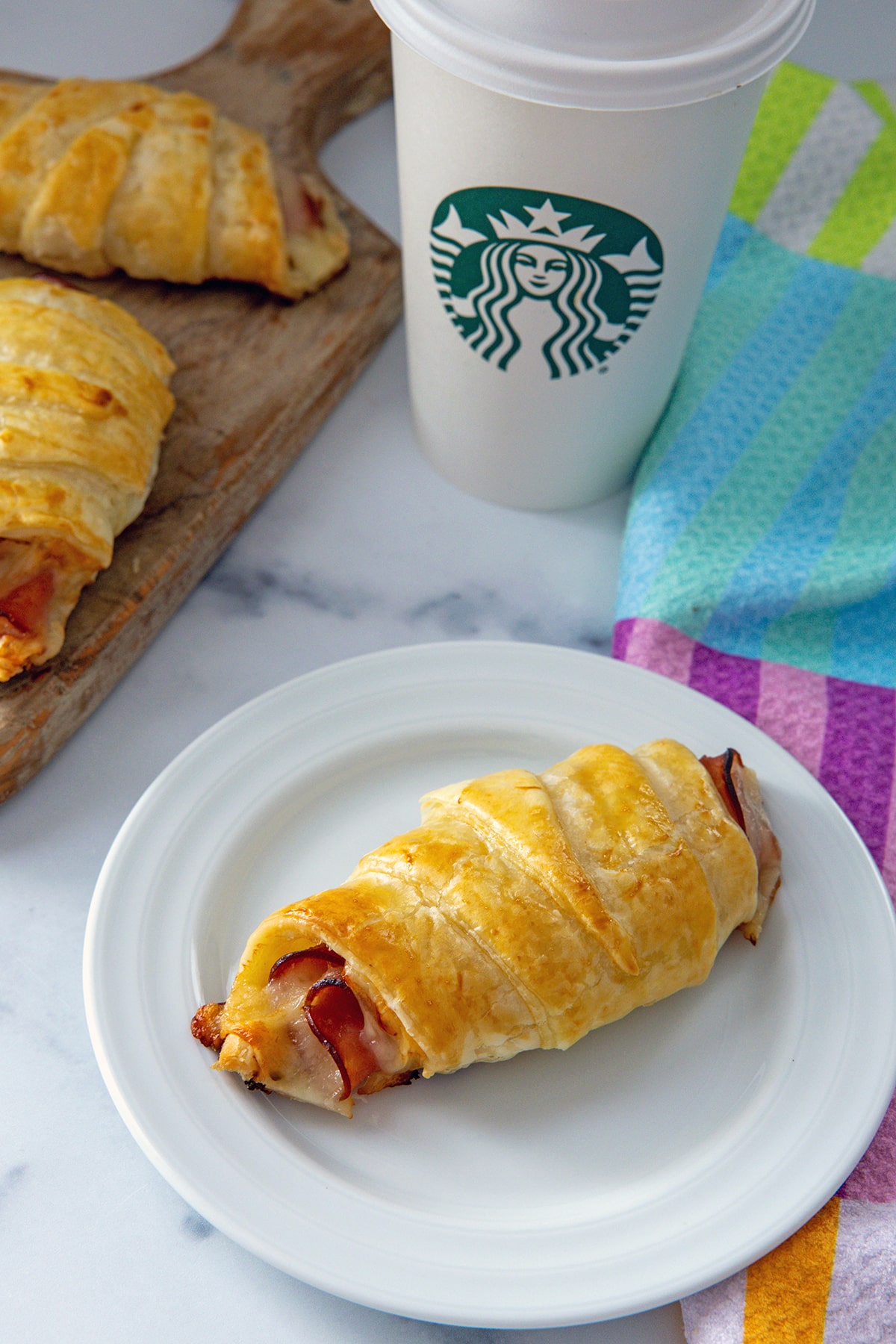 Ham and cheese croissant on a plate with Starbucks cup and more croissants in background.