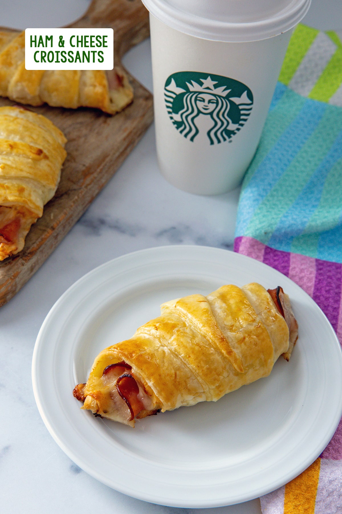 Ham and cheese croissant on a plate with Starbucks cup and more croissants in background with recipe title at top.