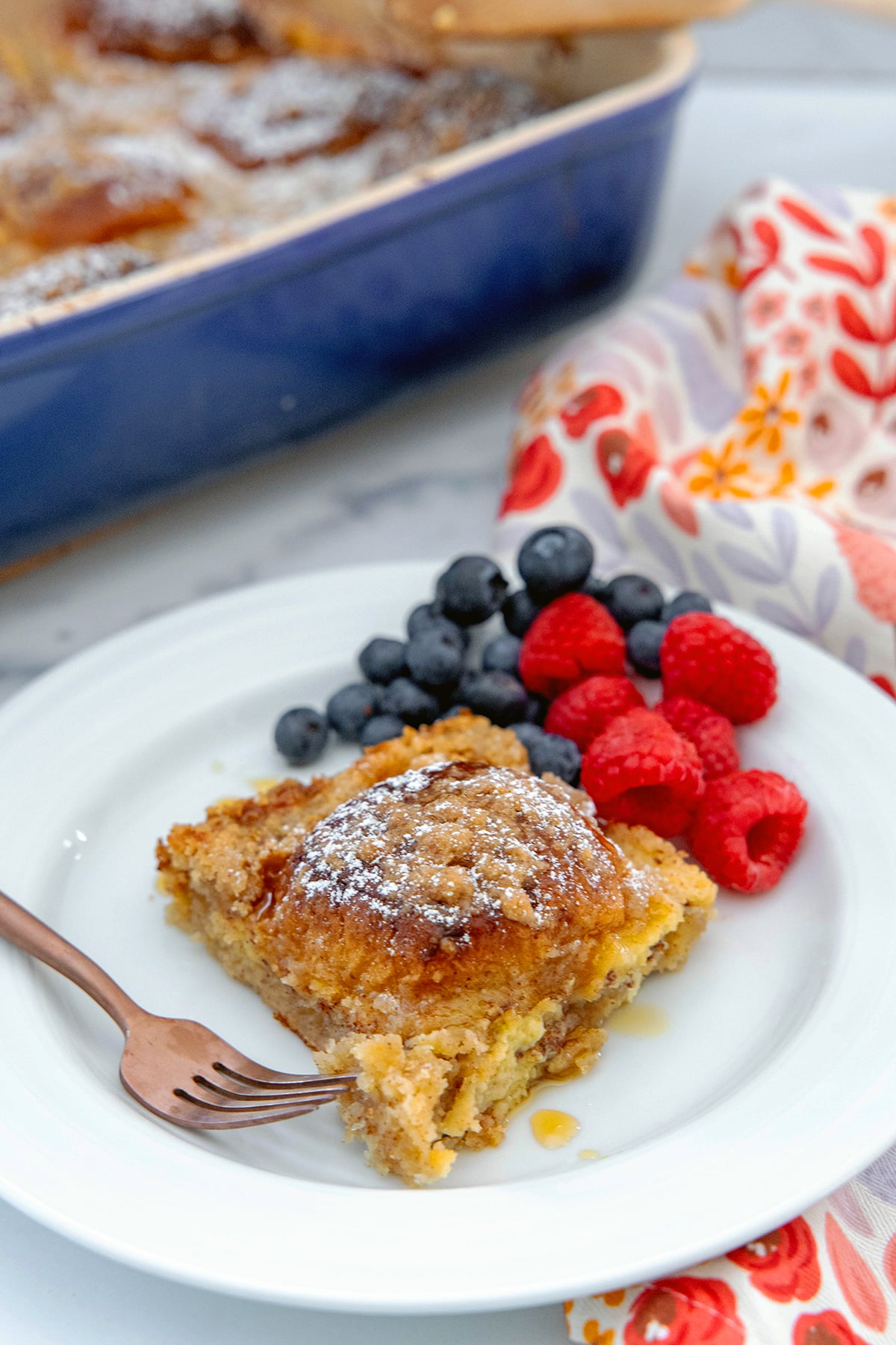 Slice of Hawaiian roll french toast casserole eon a plate with a fork and berries with casserole dish in the background.