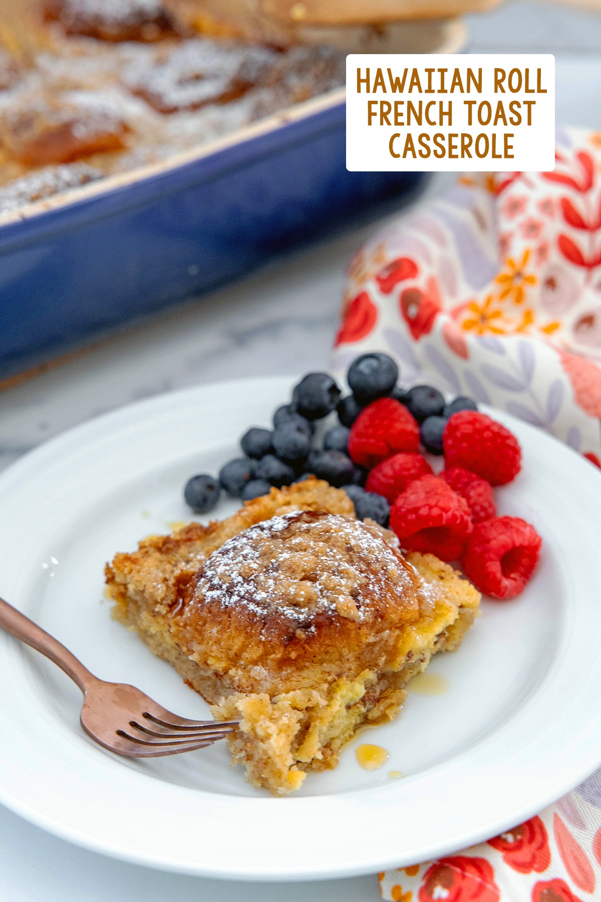 Slice of Hawaiian roll french toast casserole eon a plate with a fork and berries with casserole dish in the background and recipe title at top.