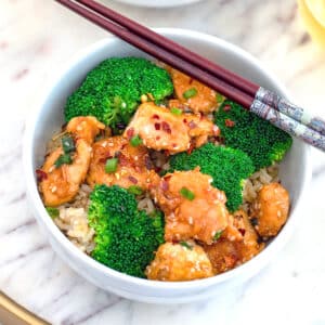 Closeup view of a bowl of healthier sesame chicken with brown rice, broccoli, and chopsticks.