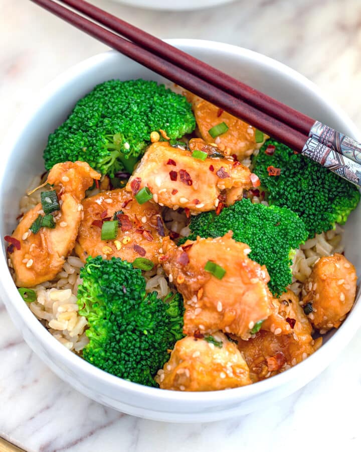 Closeup view of a bowl of healthier sesame chicken with brown rice, broccoli, and chopsticks.