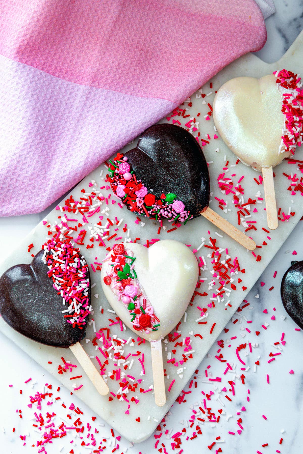 Bird's eye view of four heart cakesicles on a marble surface with lots of Valentine's Day sprinkles.