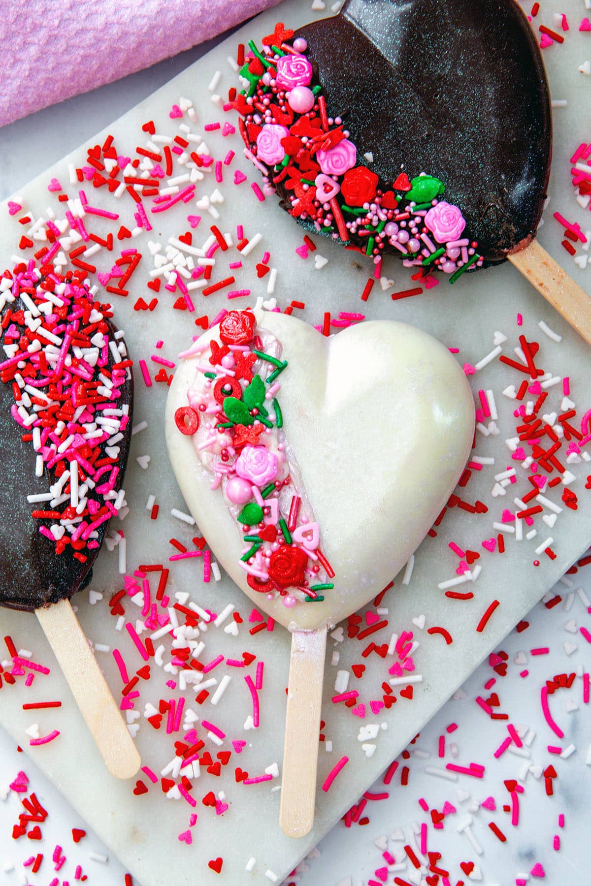 Overhead view of white chocolate and dark chocolate heart-shaped cakesicles with sprinkles all around and recipe title at top.