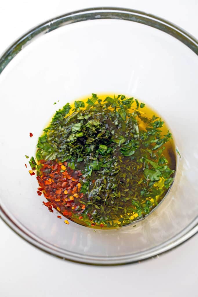 Parsley, basil, thyme, and red pepper flakes in oil in bowl.