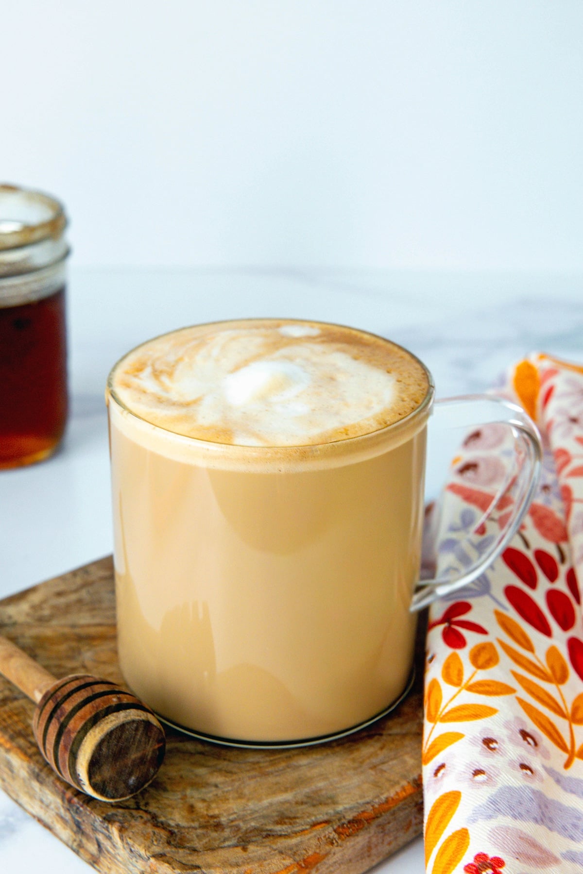 Head-on view of a honey almond milk flat white in a clear mug with honey syrup and honey stick in background.