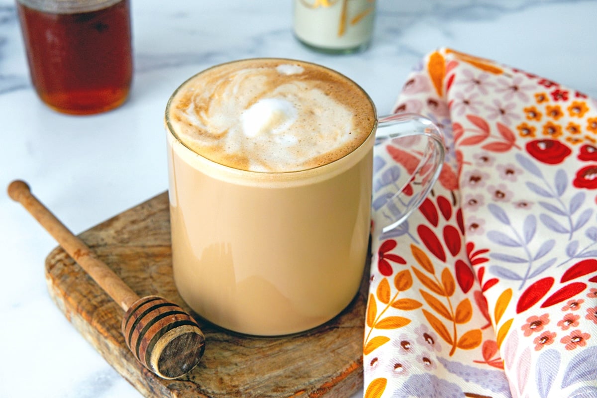 Landscape view of a clear glass mug filled with honey almond milk flat white with honey stick next to it.