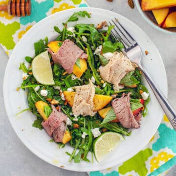 Closeup of an arugula salad with turkey and roast beef-wrapped peaches and lime wedges.