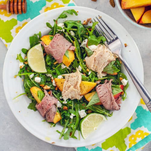 Closeup of an arugula salad with turkey and roast beef-wrapped peaches and lime wedges.