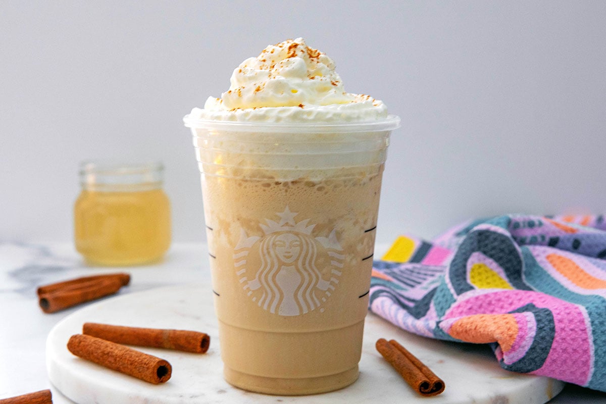 Landscape head-on view of a Horchata Frappuccino with whipped cream in a Starbucks cup with cinnamon sticks all around and jar of simple syrup in background.
