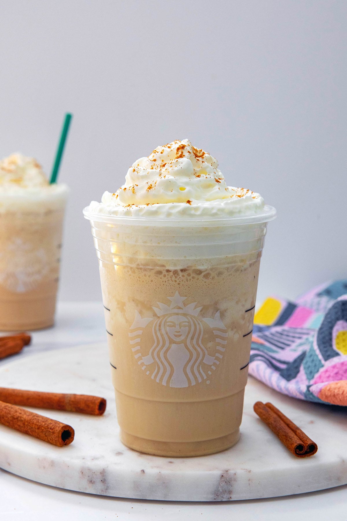 Head-on view of a Horchata Frappuccino in a Starbucks cup with cinnamon sticks around and second Frappuccino in background.